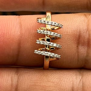 May include: A gold-toned ring with a spiral design. The ring features a series of horizontal bands encrusted with small, clear gemstones. The bands are arranged in a vertical, staggered pattern, creating a unique and eye-catching aesthetic. The ring is set against a neutral background.