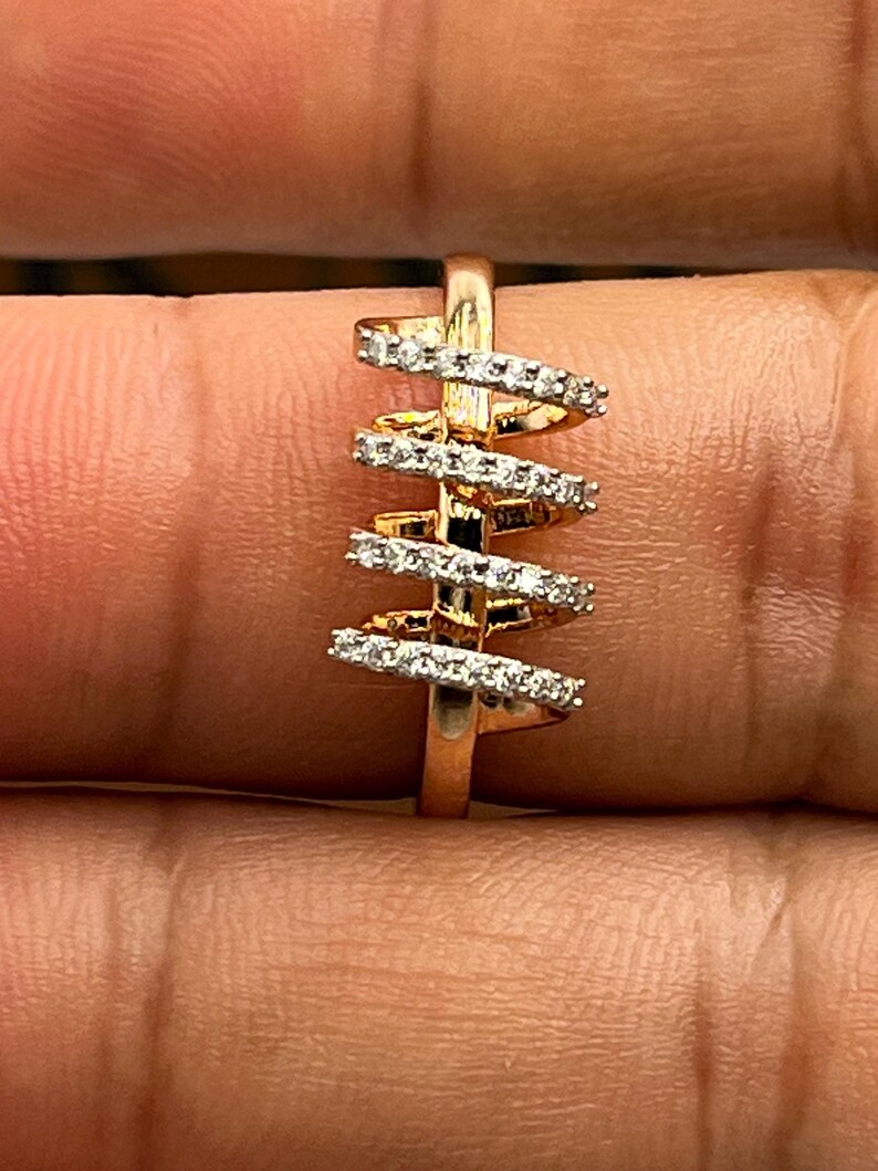 May include: A gold-toned ring with a spiral design. The ring features a series of horizontal bands encrusted with small, clear gemstones. The bands are arranged in a vertical, staggered pattern, creating a unique and eye-catching aesthetic. The ring is set against a neutral background.