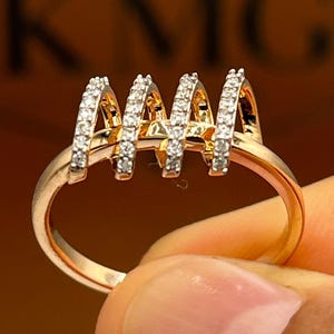May include: A gold-toned ring featuring four arched sections, each encrusted with small, sparkling stones. The ring's band is a simple, polished gold. The background is a blurred, warm tone, with the letters "KNG" visible.