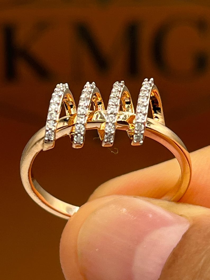 May include: A gold-toned ring featuring four arched sections, each encrusted with small, sparkling stones. The ring's band is a simple, polished gold. The background is a blurred, warm tone, with the letters "KNG" visible.