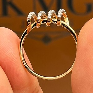 May include: A gold ring with a unique design. The ring features four arched sections, each encrusted with small, sparkling stones. The band is a simple, polished gold. The ring is held between two fingers.