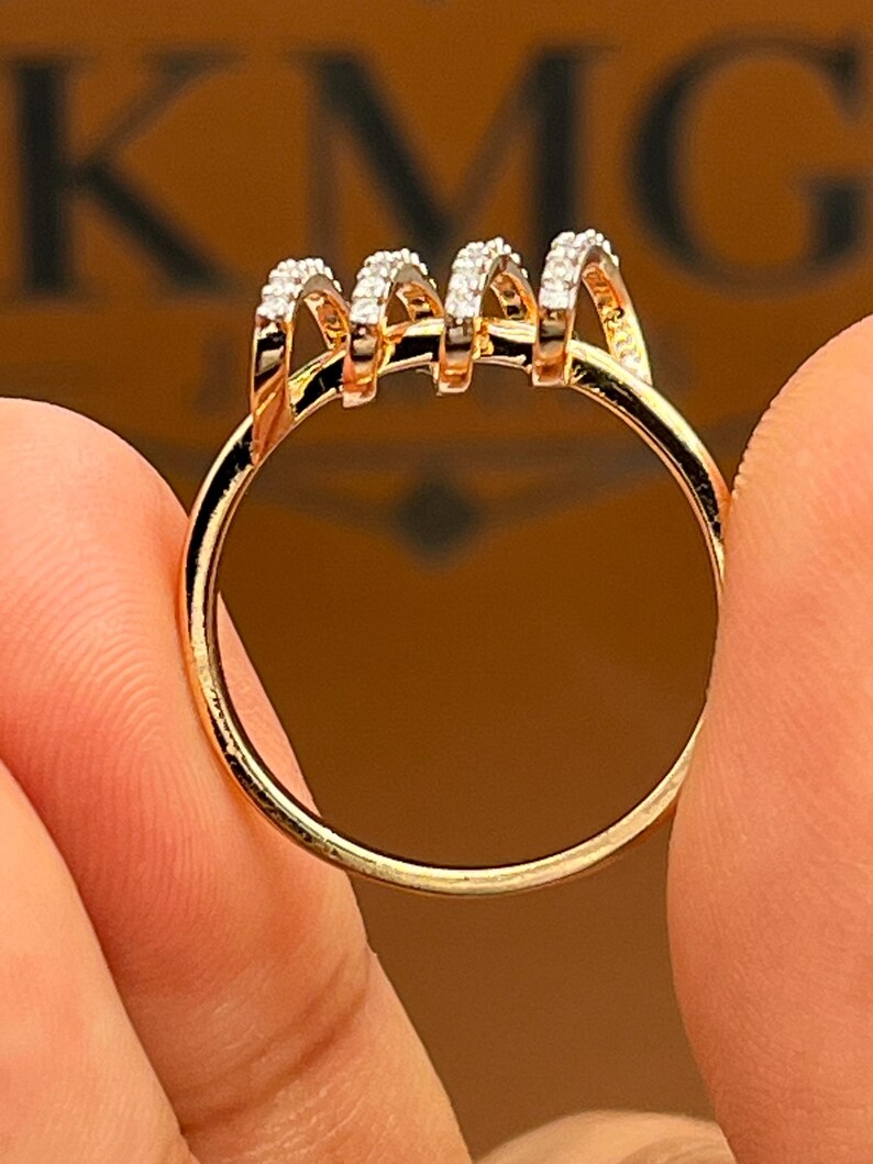 May include: A gold ring with a unique design. The ring features four arched sections, each encrusted with small, sparkling stones. The band is a simple, polished gold. The ring is held between two fingers.