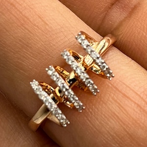 May include: A gold-colored ring with four parallel bars, each encrusted with small, clear gemstones. The ring is set against a blurred background, highlighting the intricate design and sparkle of the jewelry.
