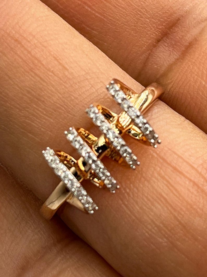 May include: A gold-colored ring with four parallel bars, each encrusted with small, clear gemstones. The ring is set against a blurred background, highlighting the intricate design and sparkle of the jewelry.