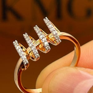 May include: A gold-toned ring with four vertical bars, each encrusted with small, sparkling white stones. The ring is held up, showcasing its design and the light reflecting off the stones. The ring's band is smooth and polished.