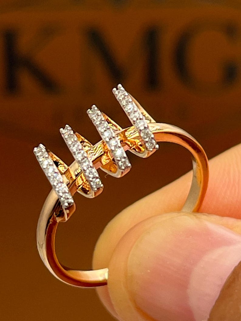 May include: A gold-toned ring with four vertical bars, each encrusted with small, sparkling white stones. The ring is held up, showcasing its design and the light reflecting off the stones. The ring's band is smooth and polished.