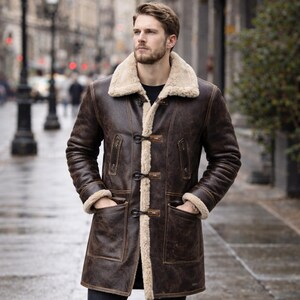 May include: A brown leather coat with a cream shearling collar and lining. The coat has two front pockets and toggle closures. The longline coat is worn by a person on a wet street. The coat's length is approximately 100 cm.