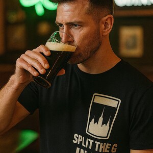 May include: A person drinking dark beer from a pint glass. The person is wearing a black t-shirt with a graphic of a pint glass and the text "SPLIT THE G IN NYC". A neon green shamrock is in the background.