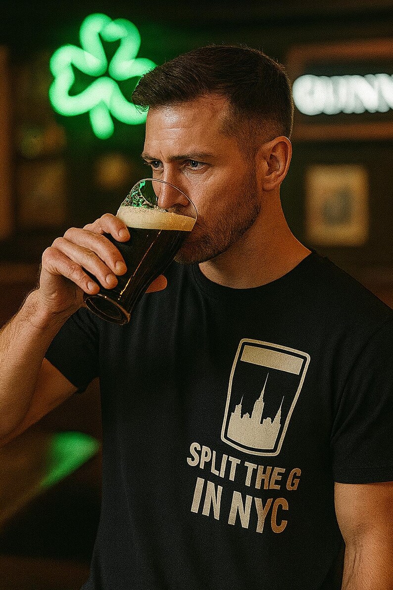 May include: A person drinking dark beer from a pint glass. The person is wearing a black t-shirt with a graphic of a pint glass and the text "SPLIT THE G IN NYC". A neon green shamrock is in the background.