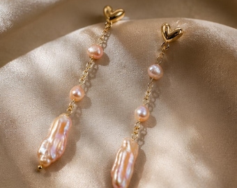 May include: A pair of gold-tone drop earrings featuring a small heart-shaped stud at the top. Each earring consists of a delicate gold chain suspending two small round pink pearls and one larger, elongated, irregular-shaped pink pearl at the base.
