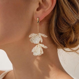 Può includere: Orecchini pendenti color oro con due pendenti bianchi traslucidi a forma di fiore. Gli orecchini hanno un design delicato, con un piccolo borchia d'oro all'orecchio e una sottile barra d'oro che collega gli elementi floreali.