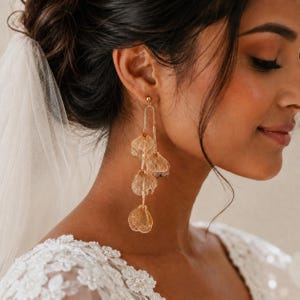 May include: A gold-toned dangle earring featuring a thin, arched metal frame from which four translucent, petal-shaped elements are suspended in a vertical chain. The earring has a delicate, layered structure with a polished metallic finish.