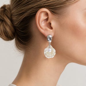 May include: A pair of silver-toned drop earrings. Each earring features a small, rounded top, a delicate chain, and a large, shell-shaped pendant with a pearl at the center. The shell pendant is a translucent, off-white color.