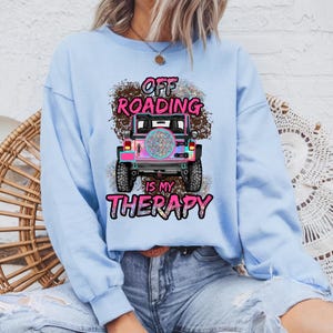 May include: Light blue sweatshirt with a graphic of a colorful off-road vehicle. The text "OFF ROADING IS MY THERAPY" is printed in pink and black. The design features a mud-like background.