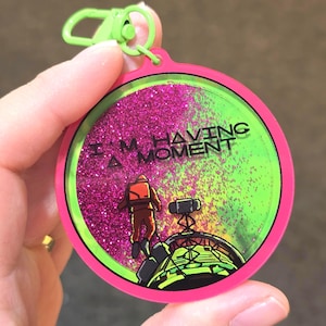 May include: A circular keychain with a bright pink border and a lime green clip. The center features a glittery pink and green background with the text I'M HAVING A MOMENT. An illustration shows a figure in an orange suit near a mechanical structure.
