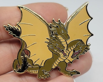 Può includere: Spilla dorata raffigurante un drago a tre teste con ali beige. Il drago ha un corpo verde e bocche aperte, con un design dettagliato. La spilla è probabilmente in metallo e ha una finitura lucida, per gli appassionati di fantasy.