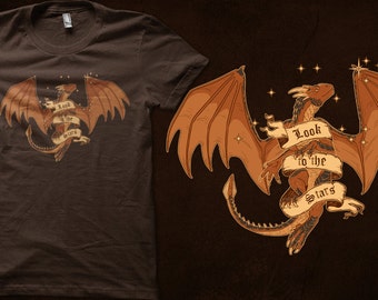 Peut inclure: T-shirt marron avec une illustration d'un dragon aux ailes orange et beige. Une bannière avec l'inscription « Look to the Stars » est présente. Le design comprend des étoiles et un thème céleste.