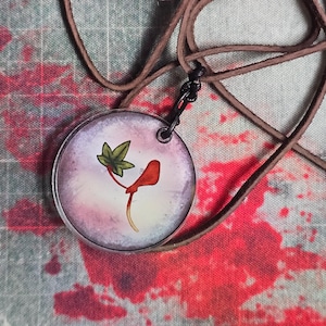 Op de afbeelding: Een ronde hangerketting met een bruin leren koord en een bedel van heldere hars. De bedel toont een gedrukte illustratie van een groen blad en een rood, gebogen botanisch element tegen een zachte, paars-witte achtergrond met kleurverloop.