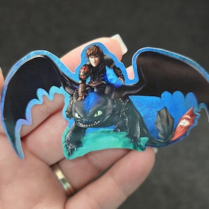 May include: A colorful sticker featuring a black dragon with large wings, ridden by a character. The dragon has green eyes and is set against a blue background. The sticker is held by a hand.