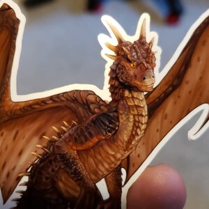 May include: A brown dragon sticker with wings spread wide. The dragon has a textured, scaly body and a golden yellow underbelly. The sticker is on a white background.