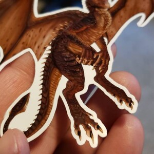 May include: A brown dragon sticker with a white outline. The dragon has wings, a long tail, and claws. The sticker is on a person's hand.