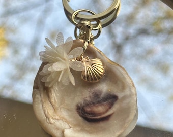 Llavero con forma de ostra, adornado con conchas marinas y flores blancas.