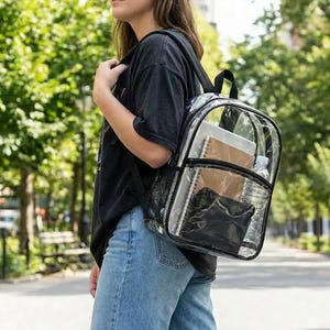 May include: Transparent backpack with black edging and straps. The clear design reveals the contents, such as notebooks and a water bottle. The backpack is being carried on a person's back.