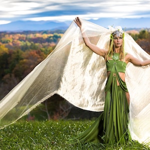 May include: A woman in a flowing green dress and headpiece stands in a grassy field, holding a large, sheer, cream-colored fabric. The background features a colorful autumn forest and mountains under a cloudy sky.