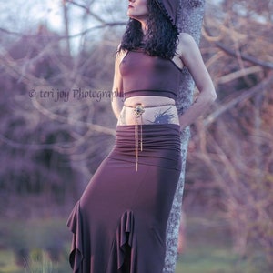 Peut inclure: Une tenue deux pi&egrave;ces violet fonc&eacute; compos&eacute;e d'un crop top et d'une longue jupe &agrave; ourlet &agrave; volants. La tenue est accessoiris&eacute;e d'une ceinture d&eacute;corative. Le mod&egrave;le porte une capuche assortie. L'arri&egrave;re-plan est une sc&egrave;ne ext&eacute;rieure floue.