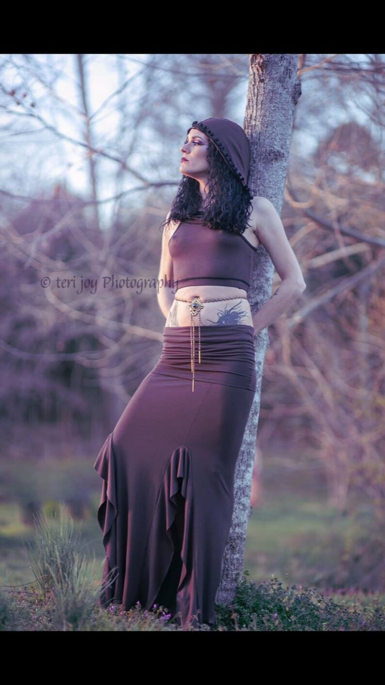 Peut inclure: Une tenue deux pi&egrave;ces violet fonc&eacute; compos&eacute;e d'un crop top et d'une longue jupe &agrave; ourlet &agrave; volants. La tenue est accessoiris&eacute;e d'une ceinture d&eacute;corative. Le mod&egrave;le porte une capuche assortie. L'arri&egrave;re-plan est une sc&egrave;ne ext&eacute;rieure floue.