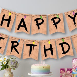 May include: A birthday party setup featuring a "Happy Birthday" banner adorned with fairy lights. A table is laden with a cake, presents, cupcakes, and a vibrant tablecloth. A vase of flowers and a jar of sweets add to the celebratory atmosphere.