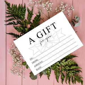 K&ouml;nnte beinhalten: Ein wei&szlig;er Geschenkgutschein mit den Worten "A GIFT for you" in schwarzer Schrift. Der Gutschein hat Zeilen f&uuml;r "TO:" "FROM:" "AMOUNT:" und "DATE:" mit einer Schleifengrafik. Der Gutschein ist von gr&uuml;nen Farnen und wei&szlig;en Blumen auf einem rosa Holzgrund umgeben.