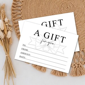 K&ouml;nnte beinhalten: Zwei wei&szlig;e Geschenkkarten mit schwarzem Text, der "A GIFT for you" lautet. Die Karten haben Zeilen f&uuml;r "TO:", "FROM:", "AMOUNT:" und "DATE:". Eine dekorative Schleifengrafik befindet sich in der Mitte. Die Karten liegen auf einem gewebten beigefarbenen Platzset.