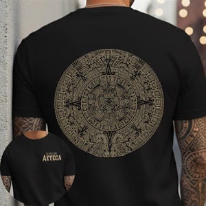 May include: Black t-shirt featuring a detailed gold-colored Aztec calendar design on the back. The shirt also has the text "SANGRE AZTECA" on the front. The design is circular and intricate, showcasing ancient cultural elements.