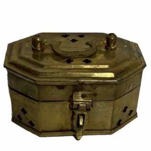Puede incluir: Una caja de latón antigua de forma octogonal. Cuenta con un asa, recortes decorativos y un cierre de pestillo. La caja tiene un acabado deslustrado, lo que sugiere edad y uso.