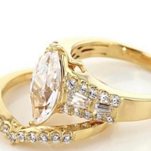 May include: A gold engagement ring set with a marquise-cut diamond and a diamond-studded band. The set includes a matching band with small, round diamonds. The rings are set against a white background.