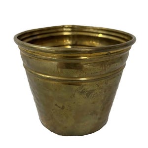 May include: A small, antique-style brass planter pot with a slightly flared rim and a series of horizontal bands. The pot has a weathered appearance, suggesting age and use. The interior is visible, and the overall design is simple and functional.
