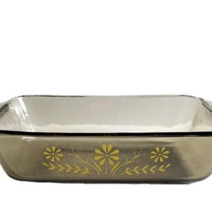 Puede incluir: Fuente rectangular de vidrio para hornear con un diseño floral. La fuente es de color marrón claro con un borde y asas negras. El diseño floral es amarillo y presenta tres flores y hojas.