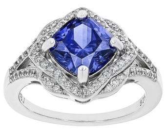 Op de afbeelding: Een zilveren ring met een grote, vierkant geslepen, diepblauwe edelsteen in het midden. De edelsteen is omgeven door kleinere, heldere accentstenen. De ring heeft een gedetailleerd, sierlijk ontwerp met een gepolijste afwerking.