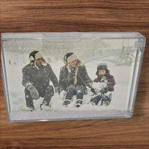 May include: A clear, rectangular snow globe with a family photo inside. The photo shows three people in winter clothing sitting in the snow. The base of the snow globe is filled with white glitter.