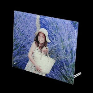 Puede incluir: Marco de fotos de vidrio con una imagen de una persona en un campo de lavanda. La persona lleva un sombrero y sostiene una bolsa de mimbre. El fondo es un campo de flores de lavanda moradas. El marco está sostenido por un soporte.