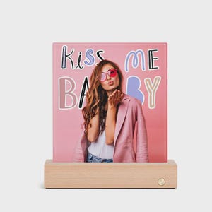 Puede incluir: Una impresión fotográfica en tonos rosas con las palabras "Kiss Me Baby" y una imagen de una persona soplando un beso, sobre una base de madera. La persona lleva una blazer rosa y gafas de sol.