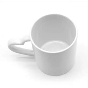 Puede incluir: Una taza de cerámica blanca con un asa en forma de corazón. La taza es cilíndrica con un acabado liso y brillante. El asa se curva con gracia, añadiendo un toque de elegancia al diseño sencillo. La taza está vacía.