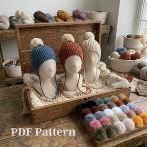 Pode incluir: Três gorros de malha canelada em azul, ferrugem e creme, cada um com um pompom a condizer, expostos em cabeças de manequim. Uma caixa de madeira contém os chapéus, com uma bandeja de pompons coloridos na frente. O texto "PDF Pattern" é visível.