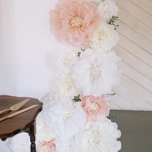 Puede incluir: Una exhibición vertical de grandes flores de papel en blanco y rosa rubor. Las flores están agrupadas, creando un telón de fondo floral. La vegetación acentúa el arreglo. Una mesa de madera con utensilios dorados para servir pasteles está en primer plano.