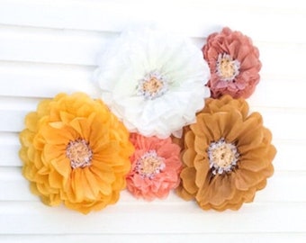 May include: A white high chair with a banner that says "ONE" in white letters on a burlap background. The banner is decorated with greenery. The chair is in front of a white wall with a large paper flower arrangement in shades of orange, yellow, and white.