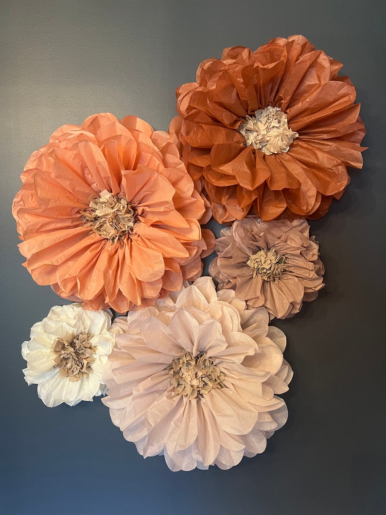 May include: Five tissue paper flowers in shades of peach, brown, and cream. The flowers are arranged in a heart shape on a dark gray background.