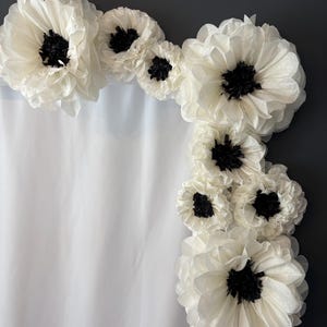 May include: A white backdrop decorated with a garland of large, white paper flowers with black centres. The flowers are arranged in a cascading pattern, creating a whimsical and elegant backdrop.