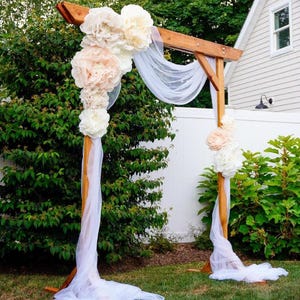 May include: A wooden wedding arch adorned with large, cream and peach paper flowers and flowing white fabric. The arch is set on a green lawn, creating a romantic outdoor setting for a ceremony.