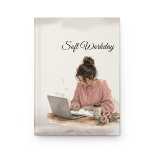 May include: A hardcover journal featuring the text Soft Workday and an illustration of a person with a brown hair bun wearing a pink sweater, sitting at a desk with a laptop, an open notebook, a pen, a coffee cup with a heart, and pink flowers.